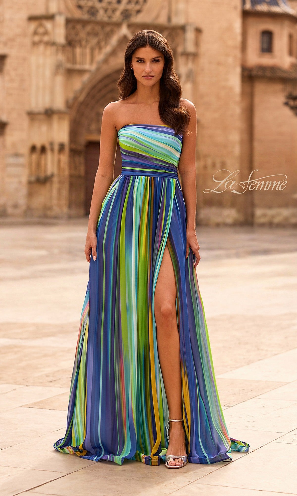 A woman stands outdoors in the La Femme 33690 multi-colored prom dress, featuring colorful vertical stripes, a strapless design, and a high leg slit, paired with white heels.